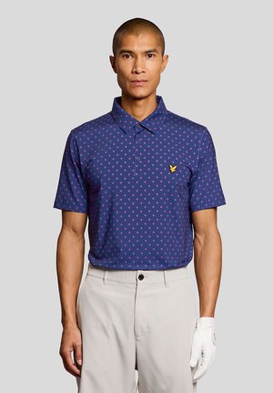 Man i marinblå piké med rosa kors, beige byxor och en vit golfhandske, stående mot en enfärgad grå bakgrund.