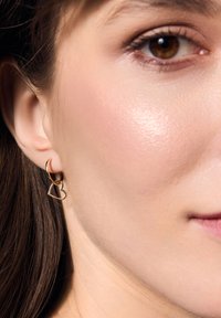 Złote kolczyki w kształcie kół z serduszkowym wisiorem. Projekt charakteryzuje się gładką fakturą i minimalistycznym stylem, podkreślając prostotę.