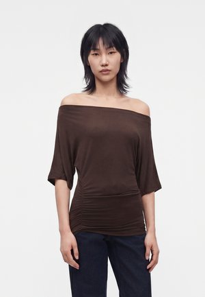 Femme aux cheveux noirs portant un haut sombre à épaules dénudées avec des côtés froncés et un pantalon sombre, debout devant un fond blanc uni.