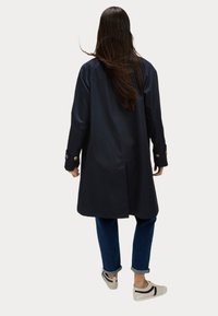 Cappotto trench navy con un taglio dritto, polsini abbottonati e un design pulito. Indossato sopra jeans blu e sneaker bianche con dettagli neri.