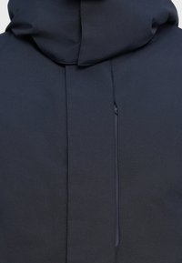 Giacca grigia scura con un ampio cappuccio strutturato, caratterizzata da un design minimalista e da una tasca con zip discreta nella parte anteriore.