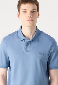 Tricou polo albastru deschis, realizat dintr-un material moale. Include o guler tradițional, o plachetă cu două butoane și logo-ul brodat "BOSS" pe piept.