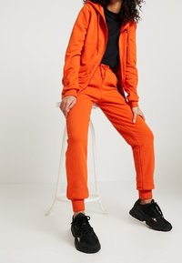 Světlý oranžový set mikiny a tepláků, obsahující zip na přední straně, stahovací pás a žebrované manžety. Nosit s černými sportovními teniskami.