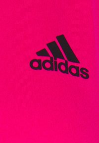 Lys pinkt stof med glat tekstur prydet med et sort Adidas-logo på venstre side, der viser tre diagonale striber over mærkenavnet.