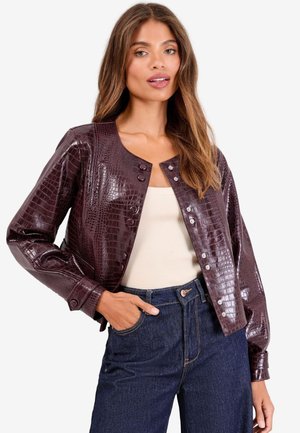 Kvinde iført en kort, mørkebrun kunstig krokodilleskindjakke over en beige top og højtaljede mørkeblå jeans, poserer med den ene hånd i lommen.