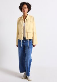 Giacca leggera giallo chiaro con tasche frontali, chiusura a scatto e orlo con cordino. Abbinata a jeans a gamba larga blu e sneaker bianche.