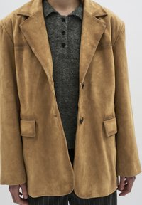 Tan suedew blazer met geknipte revers, twee voektzakken en een enkele knoopsluiting. Draag het over een grijze gebreide shirt met knopen.