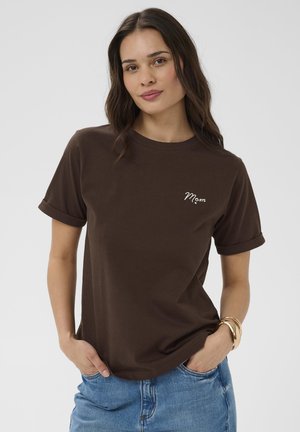 Kvinde iført en brun T-shirt med "Mom" broderet på brystet, blå jeans og guldbøjler, står med hænderne i lommerne mod en hvid baggrund.