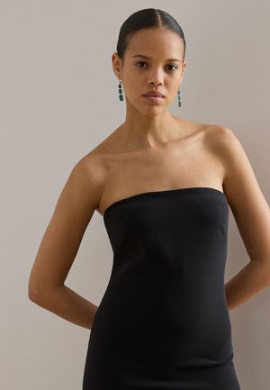 Vrouw met naar achteren gestyled haar draagt een zwarte strapless jurk en lange groene edelsteen oorbellen, staand tegen een neutrale achtergrond.