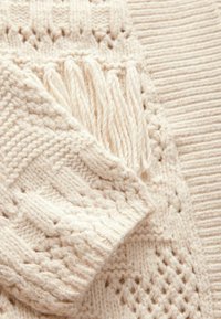 Krémový pletený svetr s texturovaným vzorem, žebrovanými manžetami a třásněmi podél okraje. Měkká, silná příze.