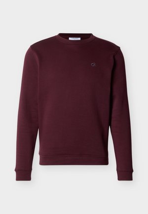 Bordo majica s okruglim izrezom i dugim rukavima te malim logotipom "ck" na lijevoj strani prsa, prikazana na jednobojnoj bijeloj podlozi.