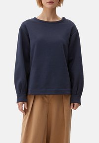 Marinblå, långärmad sweatshirt i mjukt bomull. Har en vid rund halsringning och lös passform, som kombineras med beige, högmidjade byxor.