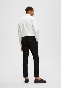 Vit skjorta med lång ärm och knapp-för krage, i kombination med slim-fit svarta byxor och mörkbruna skor. Enfärgad bakgrund.