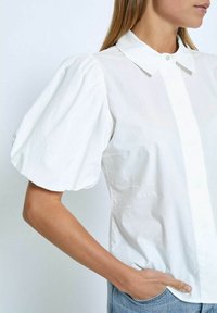 Blusa bianca in cotone con maniche a sbuffo, colletto appuntito e chiusura con bottoni. Tessuto liscio, design aderente e tasche laterali.