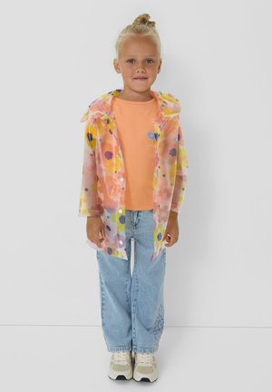 Child with blonde hair in a bun wearing a transparent floral raincoat, orange shirt, light blue jeans, and white sneakers standing against a plain background.