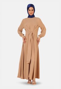 Long, beige dress with a tied waist and long sleeves, featuring a textured fabric. Paired with a blue satin hijab and light sandals.