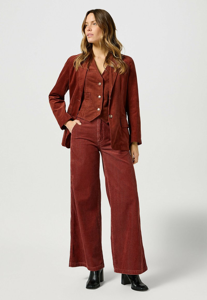 Katoenen pak in rijk bourgondisch, met een jasje met kraag en wijde broek met verticale strepen. Afgewerkt met zilveren knopen en een relaxte pasvorm.