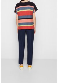 Persoon in een meerkleurig gestreept T-shirt met korte mouwen, marineblauwe broek en witte sneakers met gele hieltabs, staande met de rug naar de camera.