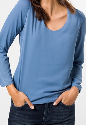 Femme portant un chemisier bleu clair à manches longues, les mains dans les poches d'un jean bleu foncé, sur un fond uni.