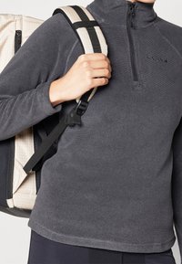 Persona vistiendo un forro polar gris oscuro con cremallera hasta la mitad, sosteniendo la correa de una mochila beige y negra sobre su hombro.