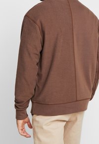 Uomo che indossa una felpa marrone a maniche lunghe e pantaloni beige, visto di spalle su uno sfondo chiaro e semplice.