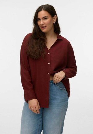 Jeune femme aux longs cheveux bruns portant une chemise bordeaux à boutons et un jean bleu clair, posant devant un fond blanc uni.