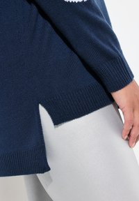 Tummansininen neulepaita, jossa on teksturoitu kangas, sivuhalkio ja ribattu hihansuu. Yhdistetty hopeisiin legginsseihin.
