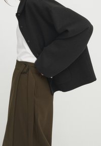 Schwarze Wolljacke mit durchgeknöpfter Vorderseite und lockerer Passform, kombiniert mit hoch taillierten braunen Hosen, die einen Gürtel und ein einzigartiges Schlaufen-Detail aufweisen.