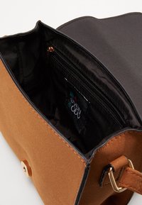 Brązowa torebka zamszowa z czarną podszewką z tkaniny. Wyposażona w kieszeń na suwak oraz złote elementy, w tym zatrzask i pasek.