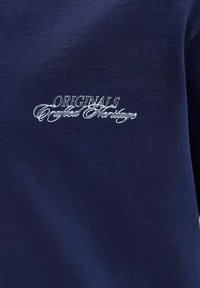 Tamsiai mėlynas medvilnės audinys su baltais siuvinėjimais tekstu "ORIGINALS Crafted Heritage", pasižymi glotnia tekstūra ir klasikinio dizaino.