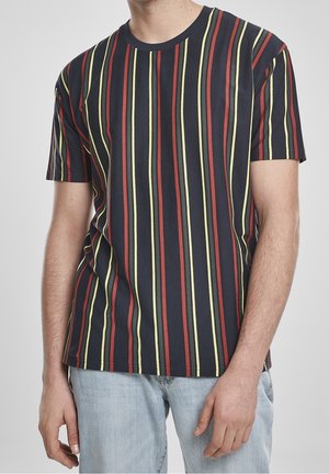 Mand iført sort T-shirt med korte ærmer med lodrette røde og gule striber og lyseblå jeans, stående mod en ensfarvet grå baggrund.