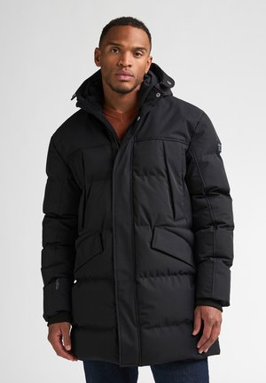 Man wearing a long black puffer jacket with a hood over a brown sweater against a plain white background.