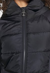 Černá páperová bunda s předním zipem, vysokým límcem a viditeln�ým šitím. Logo Barbour na táhlu zipu. Hladká textura, kompaktní design.