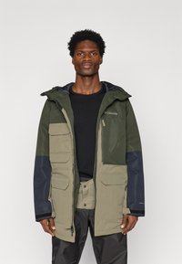 Alyvuogių žalia ir juoda nepralaidi vandeniui striukė su užtrauktuku priekyje, didelėmis kišenėmis, reguliuojama kapišonu ir kontrastingais tekstūruotais įklotais.