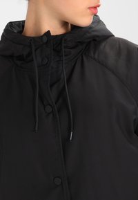 Chaqueta con capucha negra de textura suave, que presenta botones de presión, cordón y un corte holgado. Los detalles incluyen mangas raglán.