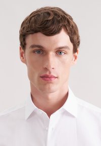 Biała koszula na guziki z klasycznym kołnierzem, o gładkiej, wyrazistej fakturze i pojedynczym guzikiem przy kołnierzu. Prosty, gładki wzór, bez żadnych zdobień.