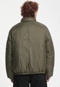 Chaqueta acolchada verde oliva con un alto cuello, dobladillo elástico y textura acolchada. Presenta un diseño trasero estilizado sin marcas visibles.