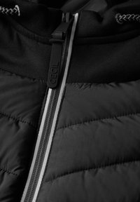 Giacca nera con zip, caratterizzata da una trama trapuntata, tessuto liscio e una cerniera argento a contrasto con un tirante etichettato "CECIL."