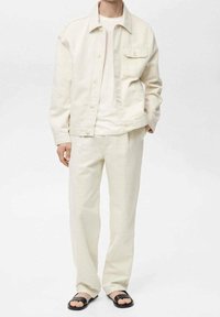 Giacca di jeans beige chiaro con una tasca, t-shirt bianca e pantaloni beige coordinati. Sandali neri completano l'outfit. Texture semplici in tutto.