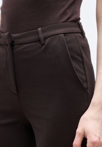 Κοντινό πλάνο ατόμου που φοράει σκούρα καφέ ραμμένα παντελόνια με θηλιές για ζώνη και πλαϊνές τσέπες, με το χέρι ορατό δίπλα στο ισχίο.