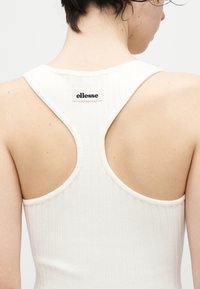 Κοντινό πλάνο ενός ατόμου που φοράει λευκό ριπ ντάνκυ τοπ με σχέδιο racerback και μια μικρή ετικέτα "ellesse" στο πάνω πίσω μέρος.