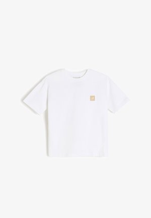 Biała koszulka z krótkim rękawem i okrągłym dekoltem, z małym beżowym kwadratowym logo po lewej stronie klatki piersiowej.