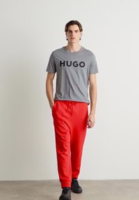 Man loopt in een grijs HUGO T-shirt, helderrode broek met koordjes en zwarte sneakers voor een witte muur.