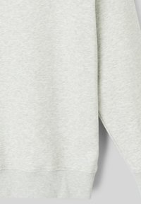 Sweatshirt gris clair en tissu doux et texturé. Présente des poignets et un ourlet côtelés, avec un subtil motif de couleur inégal et sans graphiques visibles.