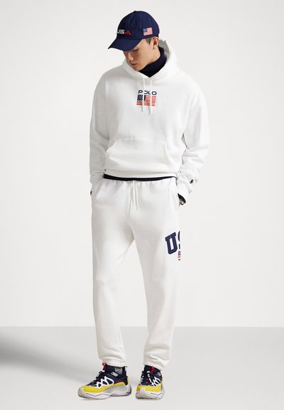 White hoodie with "POLO" and flags, paired with white sweatpants featuring "US" logo. Navy cap with logo, mixed-material athletic shoes.