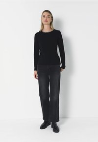 Schwarzer Pullover mit langen Ärmeln, rundem Ausschnitt und dezenten Verzierungen, kombiniert mit weiten, dunkelgrauen Jeans und schwarzen Ankle Boots.