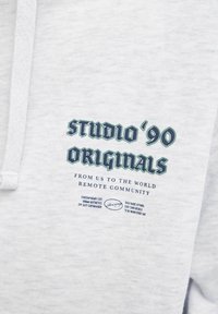 Sudadera gris claro con un diseño frontal que presenta "STUDIO '90" y "ORIGINALS" en letras verdes en negrita. Textura suave con capucha y cordón.
