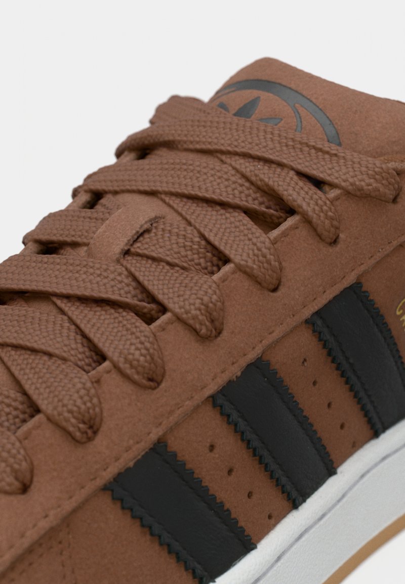 Zapatilla de ante marrón con rayas de cuero negro, diseño texturizado con cordones y suela de goma blanca. Logotipo destacado en la lengüeta.