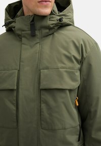 Olivovo zelená nepremokavá bunda s kapucňou, dvoma veľkými prednými vreckami a malým oranžovým akcentom na boku. Textúrovaná, ľahká látka.