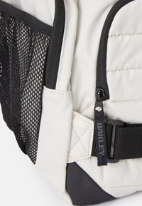 Mochila com um corpo de tecido creme, bolso em rede preto, alça preta com ferragens e etiqueta lateral com detalhe do logótipo.